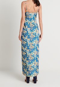 Blumenkleid mit einem blauen und gelben Muster, schmalen Trägern, enganliegendem Oberteil und fließender Silhouette. Getragen mit navyfarbenen Blockabsatzschuhen.