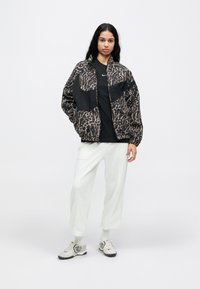 Μπουφάν bomber με τύπωμα λεοπάρ σε μαύρο και καφέ, με φερμουάρ μπροστά και ελαστικές μανσέτες. Φορεμένο πάνω από μαύρο μπλουζάκι και ανοιχτό γκρι παντελόνι με φαρδιά μπατζάκια, συνοδευόμενο από γκρι παπούτσια.