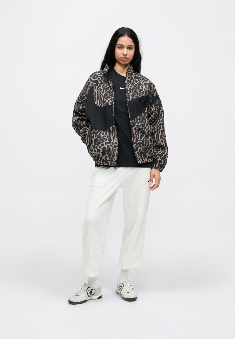 Zwart en bruin bomberjack met luipaardprint, rits aan de voorkant, elastische manchetten. Draagt over een zwart T-shirt en lichtgrijze wijde broekspijpen, met grijze schoenen.