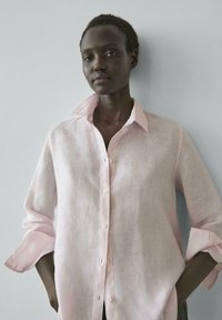 Osoba s krátkými vlasy, oblečená v světle růžové košili na knoflíky s vyhrnutými rukávy, stojící před jednobarevným světle šedým pozadím.