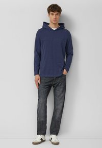 Felpa con cappuccio a maniche lunghe blu navy con tasca frontale e cappuccio, abbinata a jeans di denim scuro e sneakers beige con lacci bianchi.