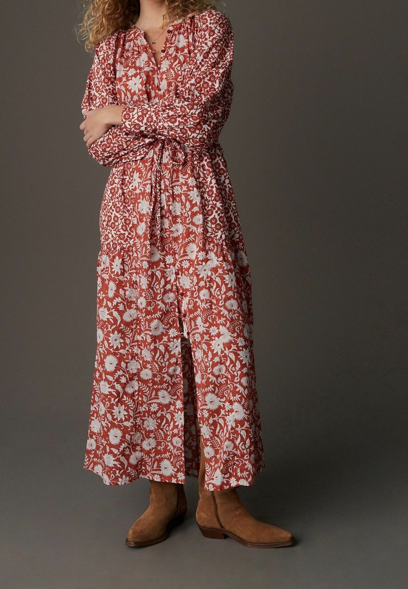 Femme portant une longue robe rouge à fleurs nouée à la taille avec des bottines en daim marron, debout les bras croisés.