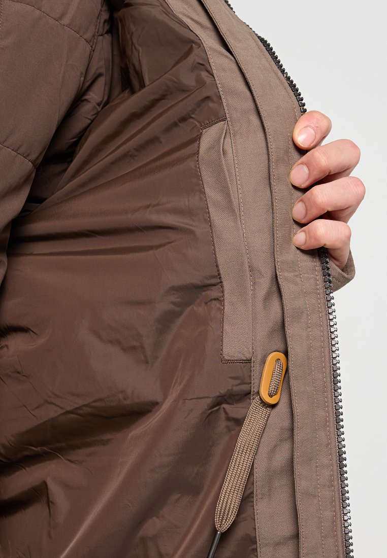 Interno della giacca isolante con un rivestimento liscio marrone e una tasca sul petto. Il materiale esterno è beige con una cerniera e un anello per la regolazione.