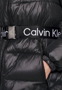 Czarna kurtka puchowa o grubym, błyszczącym tekście z szerokim paskiem Calvin Klein z białym napisem i metalową klamrą.