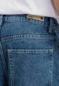 Jeans aus Denim in Hellblau, mit glatter Textur und kontrastierender Naht. Sichtbare Gesäßtaschen und ein Markenlabel aus braunem Leder, das mit "EST. 1992" gekennzeichnet ist.