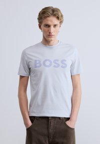 Ανοιχτό γκρι κοντομάνικη μπλούζα με τη λέξη «BOSS» σε μεγάλους μοβ χαρακτήρες. Το ύφασμα φαίνεται λείο και απαλό.