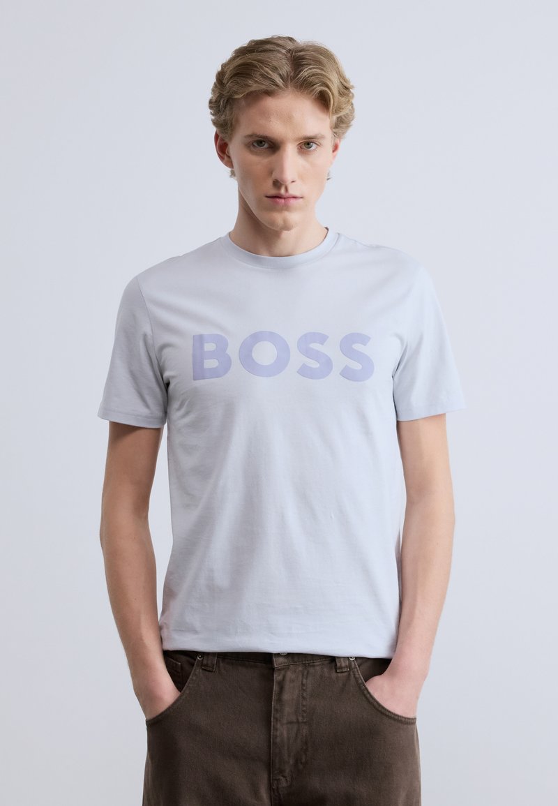 Ανοιχτό γκρι κοντομάνικη μπλούζα με τη λέξη «BOSS» σε μεγάλους μοβ χαρακτήρες. Το ύφασμα φαίνεται λείο και απαλό.