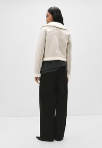 Jasnoszara kurtka ze shearlingu o skróconym kroju, z puszystym kołnierzem, kontrastującym kremowym wykończeniem i fakturowanymi ciemnymi spodniami.