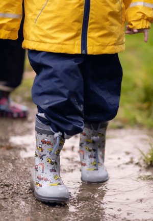 Grijze rubberen regenlaarzen met een patroon van bouwvoertuigen, gedragen met marineblauwe waterdichte broek, wandelend op een nat pad.