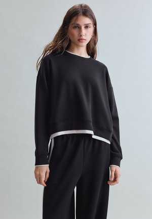 Ung kvinde med langt hår iført en sort løs sweatshirt og matchende vidde bukser mod en ensfarvet lys baggrund.