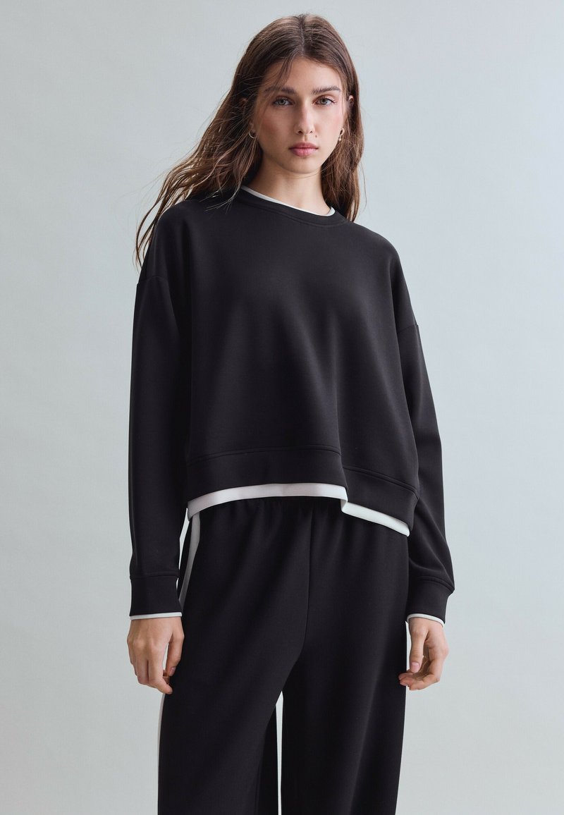 Jeune femme aux longs cheveux portant un sweat-shirt noir ample et un pantalon large assorti sur un fond clair uni.