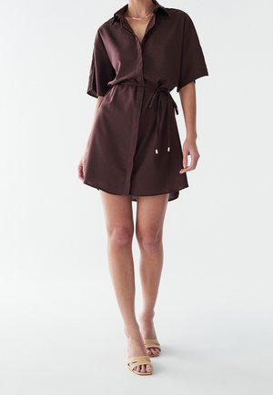 Frau trägt ein dunkelbraunes Hemdkleid mit kurzen Ärmeln und Gürtel, kombiniert mit beigen offenen Sandalen mit Absatz auf weißem Hintergrund.