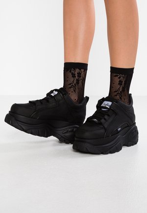Benendraag zwart grof plateau sneakers in combinatie met zwarte enkelkousen van mesh met bloemmotief op een witte vloer.