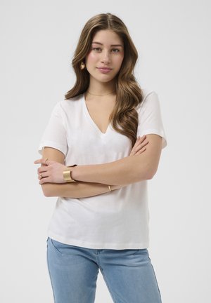Giovane donna con capelli lunghi e mossi che indossa una maglietta bianca con scollo a V e jeans azzurri chiari, in piedi con le braccia incrociate su uno sfondo uniforme.