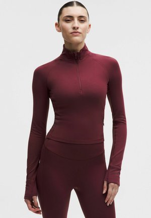 Vínovočervené športové tričko s dlhým rukávom, vysokým golierom a štvrťovým zipsom. Vyrobené z hladkej, pružnej látky. Má otvory na palce na manžetách.