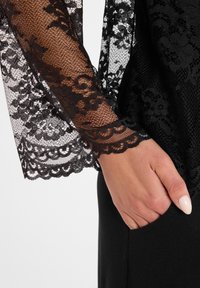 Zwarte kanttop met lange mouwen, voorzien van bloemenpatronen en een golfrand, gecombineerd met een effen zwart kledingstuk en een verborgen zak.