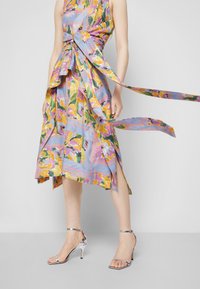 Robe imprimée de fleurs avec un fond bleu clair et des motifs roses, jaunes et verts ; tissu noué à la taille ; fente sur le côté. Talons argentés.