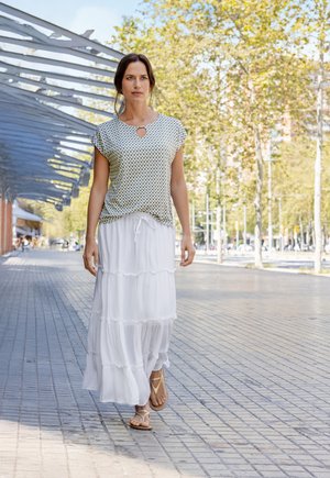 Top verde a maniche corte con un motivo a fantasia e dettaglio a chiave, abbinato a una gonna maxi bianca a balze e sandali piatti beige, su un marciapiede pavimentato.