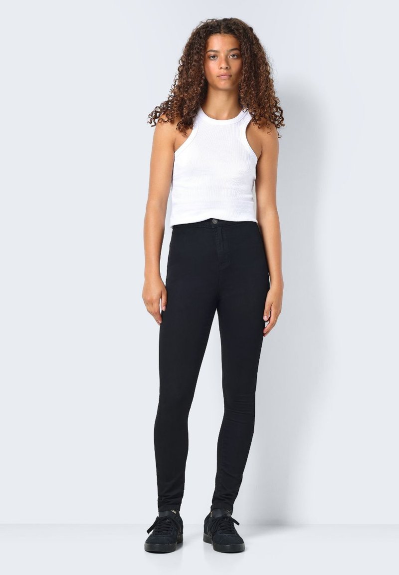 Jeans skinny pretos de cintura alta, combinados com uma t-shirt branca de malha. Modelo em pé, a usar sapatilhas pretas. Design simples.