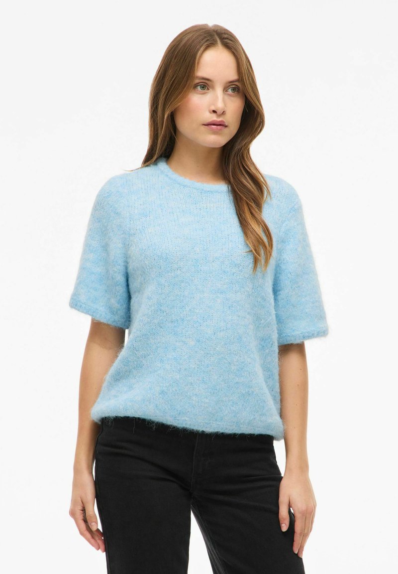 Lys blå genser med korte ermer i et mykt, loddet materiale. Rund hals og avslappet passform, kombinert med svarte bukser. Enkel og koselig design.