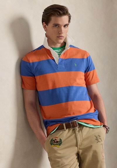 Ανδρικό πόλο μπλουζάκι με έντονες οριζόντιες ρίγες σε πορτοκαλί και μπλε, με λευκό γιακά. Συνδυάζεται με χακί παντελόνια διακοσμημένα με κεντήματα.