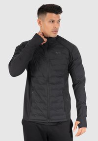 Veste isolée noire avec un devant matelassé, des manches lisses, un col haut et des poches zippées. Comprend des ouvertures pour les pouces dans les poignets.