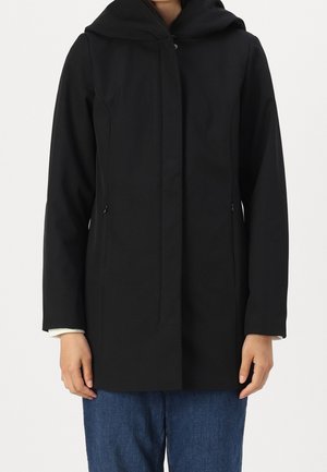 Personne portant un manteau à capuche noir à manches longues et un jean bleu, se tenant devant un fond clair uni.