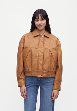 Junge Frau mit glatten dunklen Haaren, die eine beige Lederjacke und blaue Jeans trägt, steht vor einem einfarbigen hellen Hintergrund.