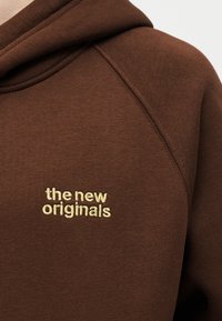 Brauner Hoodie mit beigem, auf der Brust gesticktem Text "the new originals", zeigt Stoffstruktur und Details der Schulternaht.
