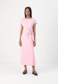 Rosa, knelengde kjole laget av mykt stoff, med korte ermer og en frontknute detalj. Modellen har på seg svarte leiligheter.