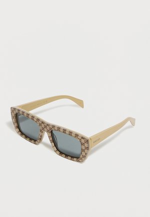 Beige rechteckige Gucci-Sonnenbrille mit dunklen Gläsern und GG-Muster auf dem Rahmen, Gucci-Logo an den Bügeln, vor weißem Hintergrund.