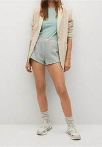 Beige Blazer über einem hellgrünen Rippoberteil, kombiniert mit grauen Shorts. Trägt weiße Sneaker und graue gestreifte Socken. Einfaches Design, lässiges Outfit.