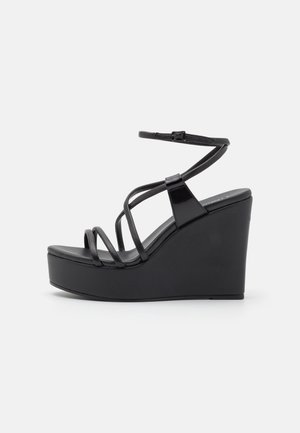 Sandales compensées noires avec des brides en cuir verni brillant, une plateforme empilée et une silhouette lisse et courbée avec une fermeture à boucle.