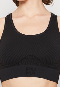 Gros plan sur une personne portant un soutien-gorge de sport noir Puma avec un tissu texturé et des panneaux en maille sous la poitrine.