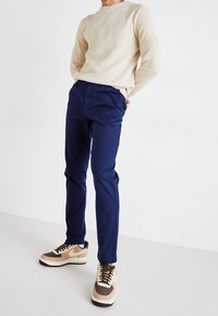 Mörkblå byxor med slim fit, kombinerat med en strukturerad beige tröja och bruna sneakers med gula detaljer och vita inslag.