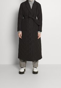 Dlouhý černý prošívaný kabát s páskem v pase, dvěma předními kapsami a s véčkovým límcem, vrstvený přes šedý rolák a kostkované kalhoty.