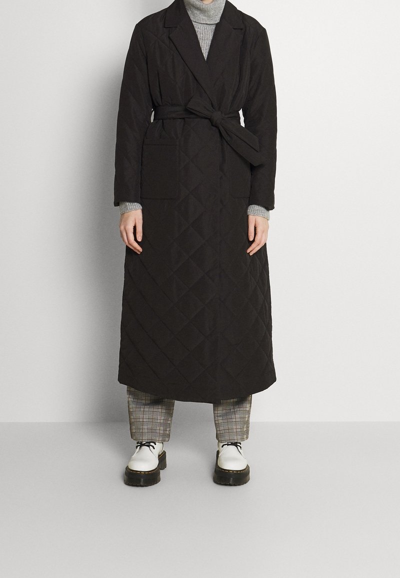 Dlouhý černý prošívaný kabát s páskem v pase, dvěma předními kapsami a s véčkovým límcem, vrstvený přes šedý rolák a kostkované kalhoty.