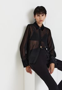 Camicia nera trasparente con davanti abbottonato e colletto, con polsini neri opachi a contrasto. Abbinata a pantaloni aderenti neri eleganti.