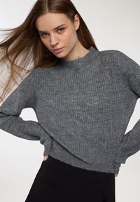 Młoda kobieta z długimi brązowymi włosami, ubrana w szary sweter dzianinowy i czarne spodnie, pozująca z rękami na biodrach na białym tle.