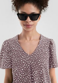 Blusa viola con motivo a pois bianchi, scollatura a V, maniche corte a sbuffo, abbinata a occhiali da sole neri angolari. Tessuto dalla consistenza liscia.