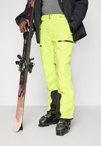 Άτομο που φορά φωτεινά κίτρινα σκι παντελόνια και μαύρα σκι μποτάκια, κρατώντας ένα ζευγάρι καφέ και μαύρων σκι.