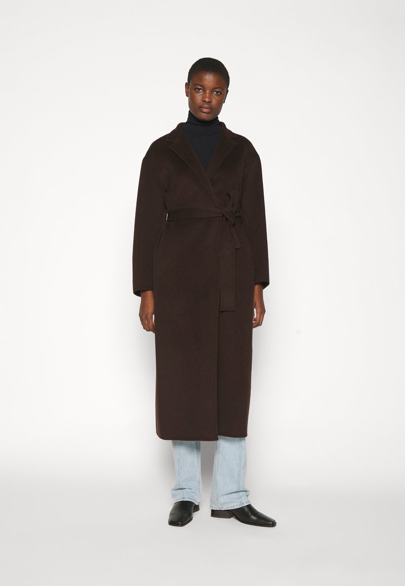 Brun uldblandet lang frakke med sjalkrave, bælte i taljen og lange ærmer, parret med lyseblå denim og sorte sko.