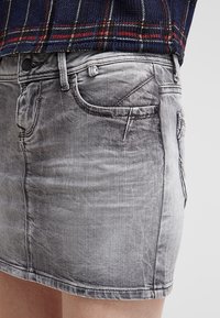 Šedá džínsová mini sukně s klasickým pětivreckovým designem, jemným ošoupáním a třepeným lemem. Materiál má hladkou, mírn�ě vybledlou texturu.