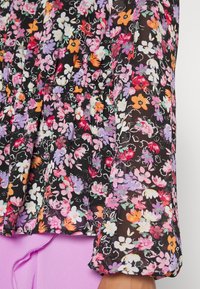 Blouse fleurie avec un fond noir orné de fleurs violettes, roses et orange vibrantes, taille élastique froncée et manches longues.