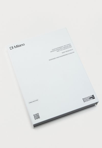 Άσπρο σκληρόδετο βιβλίο με τίτλο "DI Milano" με κείμενο για τον μονοχρωματικό μινιμαλισμό, την ενδελεχή έρευνα και το σχεδιασμό του Μιλάνου σε λευκό φόντο.