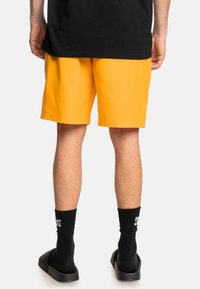 Gelbe Shorts aus leichtem Stoff, mit geradem Schnitt und Gummizug. Tragen mit schwarzen Socken und Schlippersandalen.