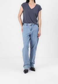 Žena v tmavě šedém krátkém rukávu s výstřihem do V, světlých volných džínách a černých botách s špičatou špičkou a sponami, stojící na bílém pozadí.