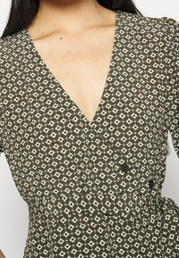 Hnědé šaty s vzorem, které mají hluboký výstřih do V, krátké rukávy a zavázaný pas se dvěma knoflíky. Tkanina má texturovaný, geometrický design.