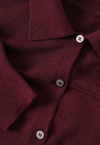 Cardigan en maille rouge foncé avec col replié et trois boutons blancs visibles sur le devant.