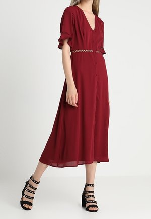 Frau trägt ein tiefrotes Midikleid mit flatternden Ärmeln und Knopfleiste vorne, kombiniert mit schwarzen, mit Nieten besetzten High-Heel-Sandalen, steht an einer weißen Wand.